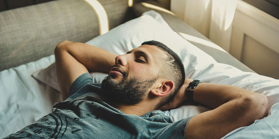 a man with beard wearing a grey t-shirt and a watch with black strap, sleeping soundly on a brown sofa bed and white pillow